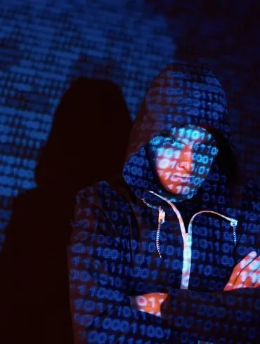 Hooded figure at a laptop representing a cybercriminal
